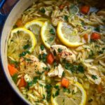 Overhead shot of a pot of chicken orzo soup with lemon slices, carrots, and herbs.