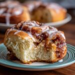 Close-up of a glazed cinnamon roll on a plate, showing a bite taken out.