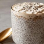 A glass filled with Coconut Chia Pudding Recipe, topped with cream and chia seeds, next to a golden spoon.