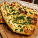 Close-up of Garlic Cottage Cheese Naan, golden brown and topped with fresh herbs, on a wooden board.