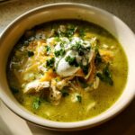 A bowl of Green Enchilada Chicken Soup topped with shredded cheese, sour cream, and fresh cilantro.
