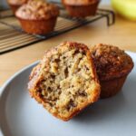 Inside view of a Healthy Banana Muffin showing the moist crumb texture. Another muffin sits whole on a plate.