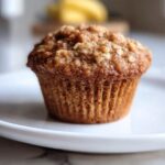 A single healthy banana oatmeal muffin sits on a white plate, showcasing its golden-brown color and oat topping.