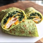 A sliced Keto Spinach Wrap showing layers of spinach, cheese, turkey, and bacon inside a green spinach tortilla.