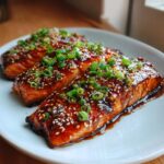 Three glazed Mango Teriyaki Salmon fillets on a white plate, garnished with sesame seeds and green onions.