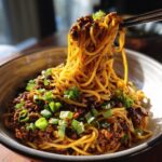 A bowl of delicious Mongolian Ground Beef Noodles topped with fresh chopped green onions, being lifted with chopsticks.