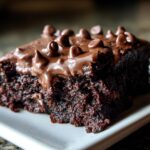 A decadent slice of Secretly Healthy Zucchini Brownies with chocolate frosting and chocolate chips on a white plate.