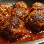 Close-up of several tender red wine meatballs simmering in a rich, red sauce in a pan.