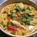 Close-up of a bowl of Thai Coconut Chicken Soup with chicken, vegetables, cilantro and a lime wedge.