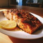 A beautifully Grilled Salmon fillet served with a lemon wedge on a white plate.
