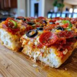 Close-up of a slice of Monster French Bread Pizza with pepperoni, olives, and green peppers, cheese pull.