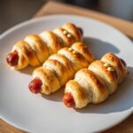 Three Mummy Pigs in a Blanket, baked golden brown, on a white plate with mustard 'eyes'.