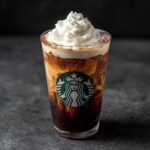 A Starbucks drink 2025 with layers of coffee and cream, topped with whipped cream, against a dark background.