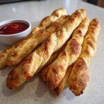 Several twisted Witch-Finger Breadsticks piled together with a side of red dipping sauce.