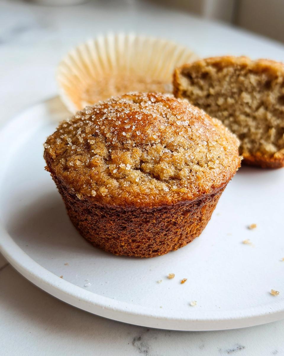 A close-up of a moist Bakery Style Banana Muffins topped with coarse sugar, with one muffin cut in half behind it.