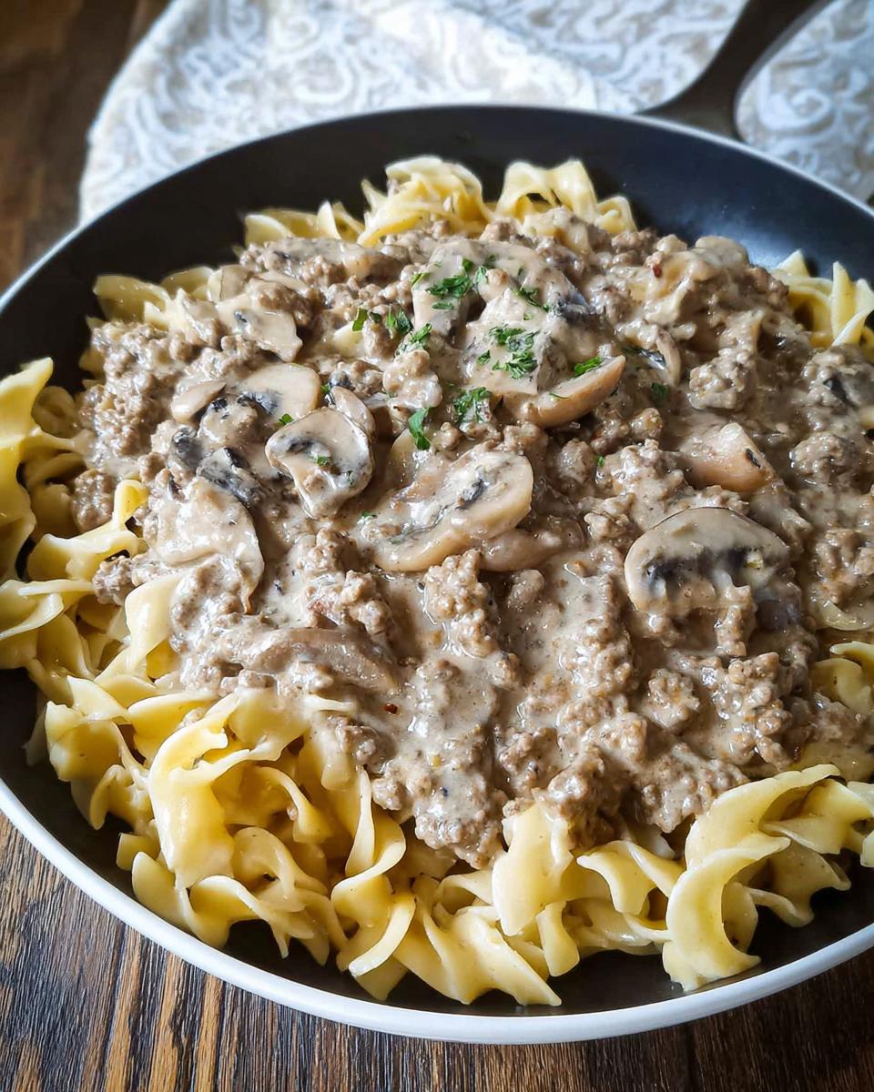 A close-up of creamy Ground Beef Stroganoff served over wide egg noodles in a dark skillet.