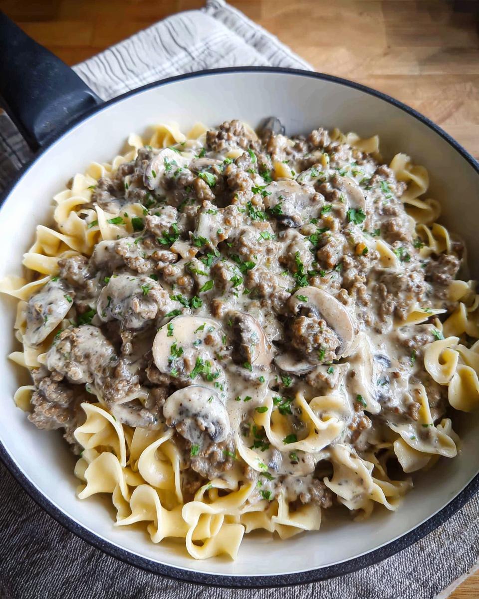 Close-up of creamy Ground Beef Stroganoff with mushrooms and parsley over egg noodles in a white skillet.