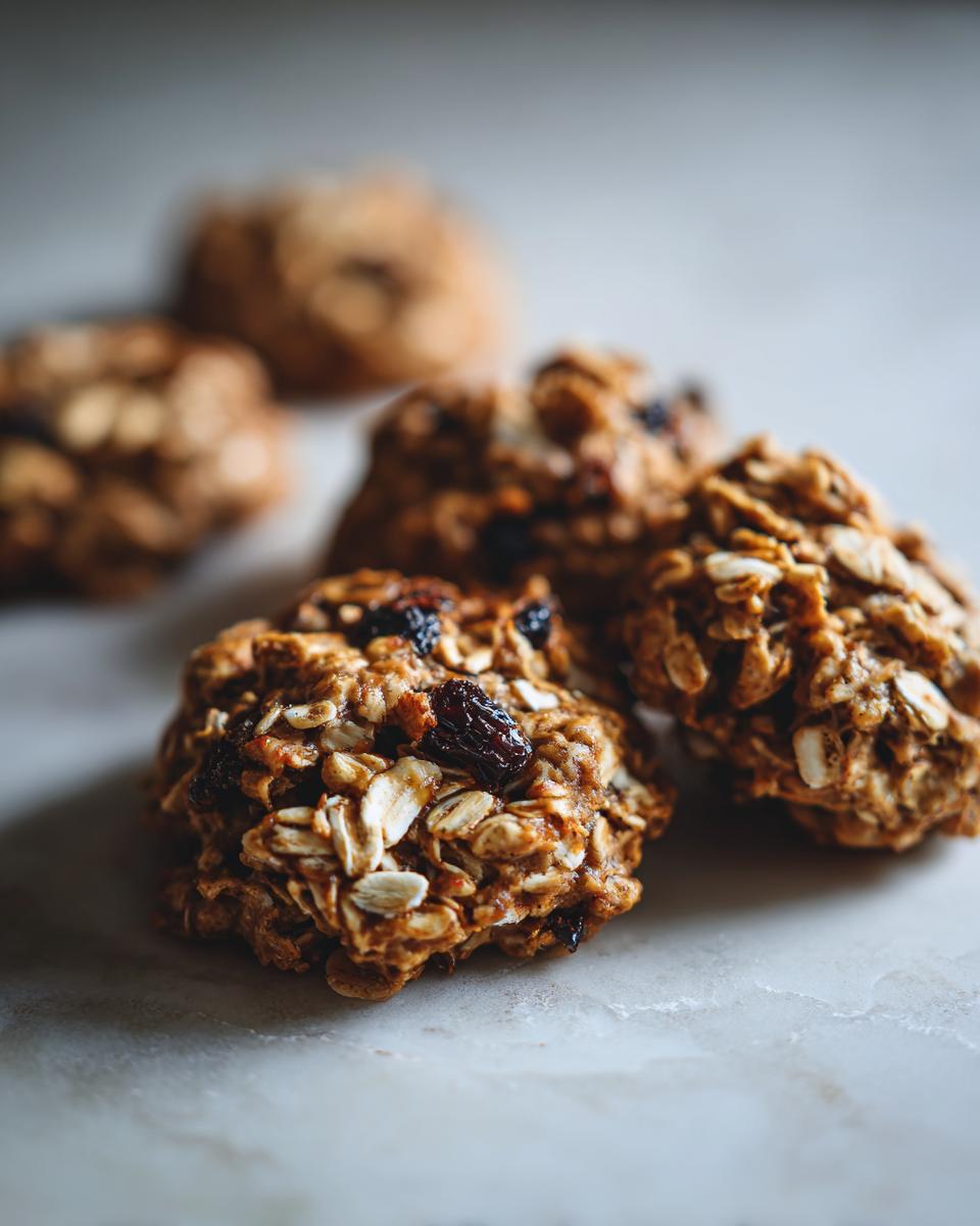 Close-up of several textured Healthy Banana Oatmeal Cookies studded with visible rolled oats and dark raisins on a light marble surface.