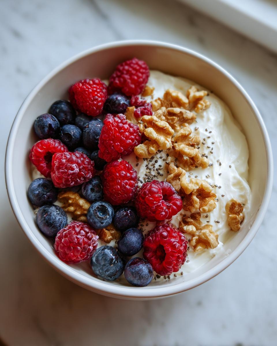 A close-up overhead shot of a High-Protein Greek Yogurt Breakfast Bowl topped with fresh raspberries, blueberries, walnuts, and chia seeds.