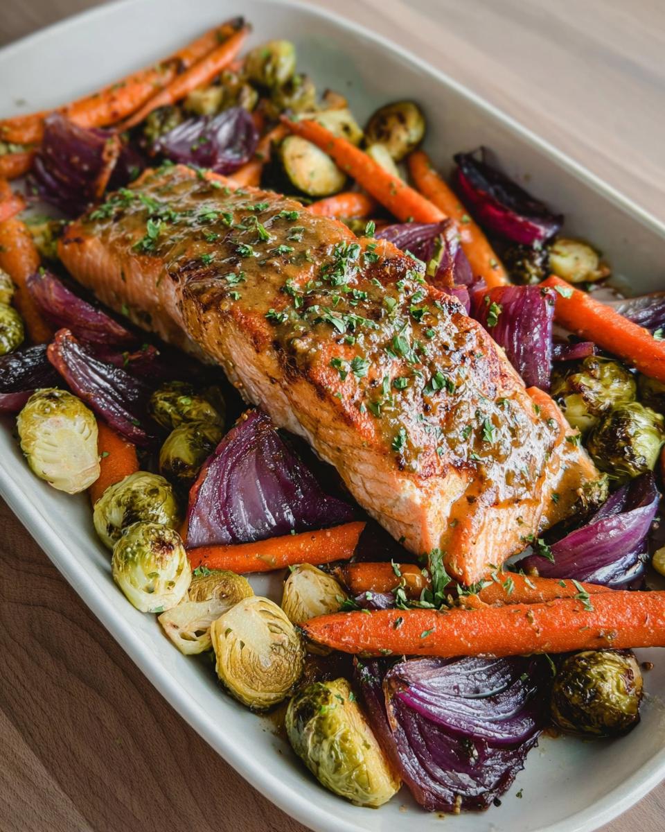 A fillet of Maple Dijon Salmon Sheet Pan glazed and topped with herbs, surrounded by roasted carrots, red onion, and Brussels sprouts.