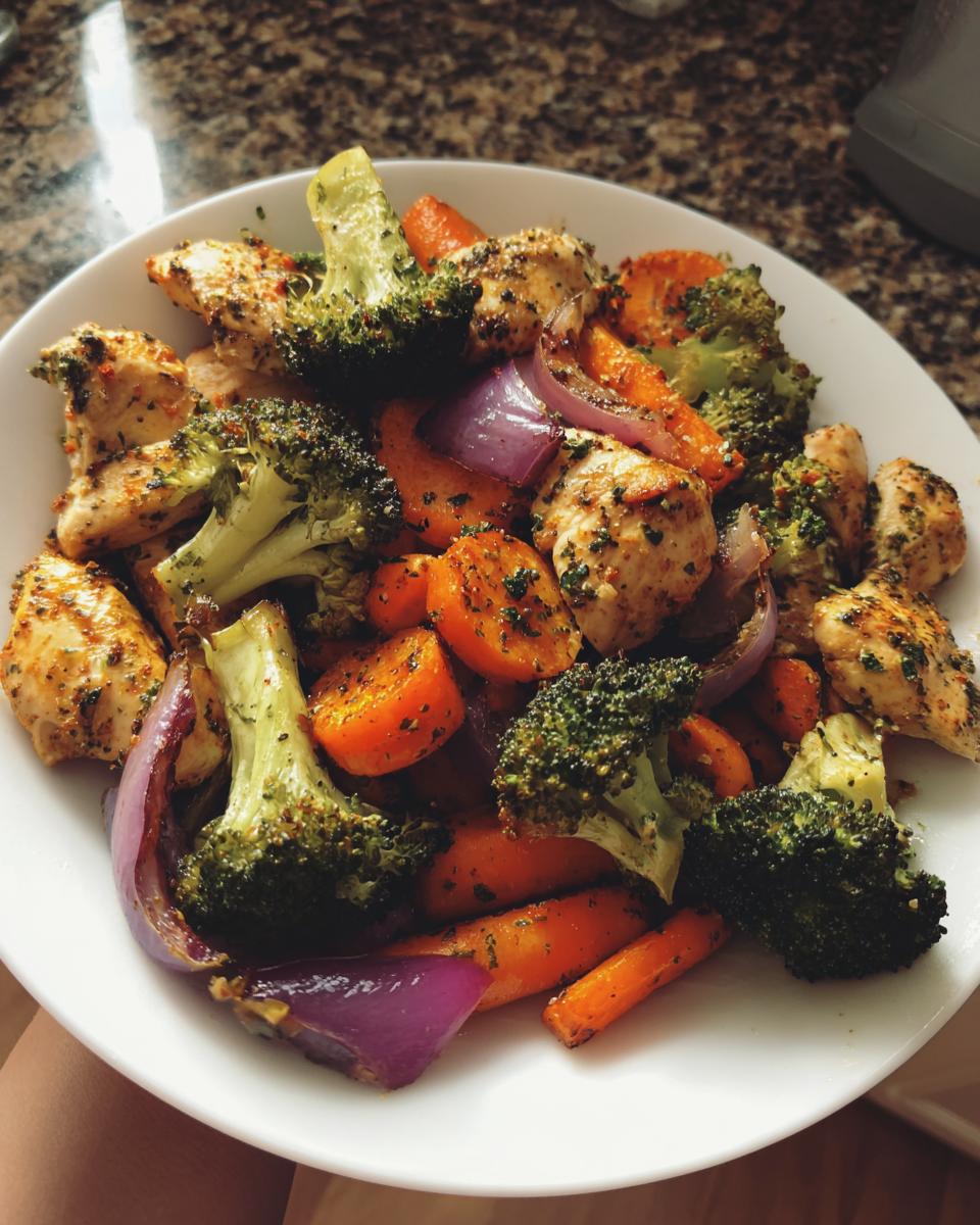 A white bowl filled with seasoned chicken pieces, roasted broccoli, carrots, and red onion from a Sheet Pan Chicken & Veggie Dinner.