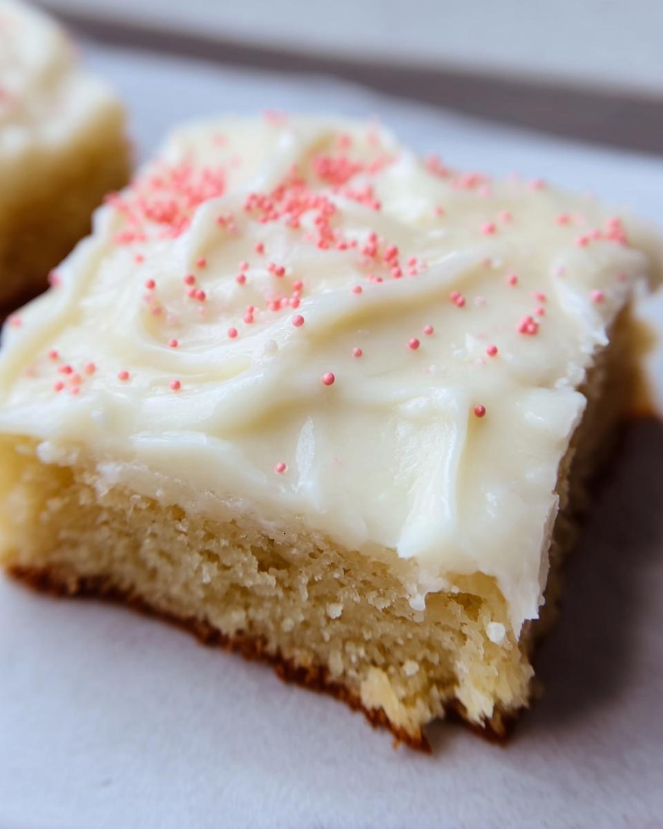 A close-up of a square Almond Flour Sugar Cookie Bars topped with thick white frosting and pink sprinkles.
