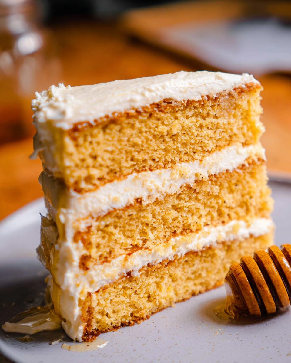 A tall slice of Layered Cornbread Cake with three layers of cake and white frosting, served with a honey dipper.